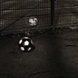 Schwarzweißaufnahme. Drei Kinder klettern über den hohen Zaun vor einem Hartplatz, vor und hinter dem Zaun liegt jeweils ein Fußball