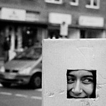 Schwarzweißaufnahme: Eine junge Frau an einer Straßenkreuzung, ihr Kopf ist von einem würfelförmigen Karton bedeckt, in den ein quadratisches Loch geschnitten ist, durch das sie lächelnd hindurchsieht.