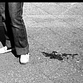 Schwarzweißaufnahme: Ein Bürgersteig. Angeschnitten am oberen Bildrand ein Kind stehend auf einem Kinderwagen, daneben die Füße der Mutter, danaben ein Fleck (Blut?), im Vordergrund der Schatten des Fotografen.